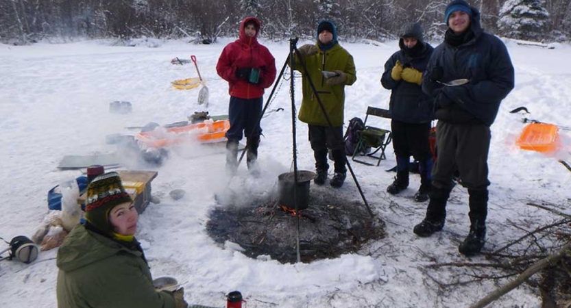 A group of people wearing winter gear stand in a snowy landscape around a campfire 